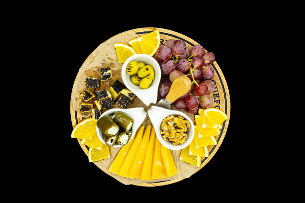 A top-down view of an assorted cheese and fruit platter arranged on a rustic wooden board. The spread includes sliced yellow cheese, grapes, orange wedges, olives, stuffed peppers, walnuts, and cubed cheese with a dark coating. The composition is colorful, symmetrical, and isolated on a black background, making it suitable for food, catering, hospitality, healthy eating, entertaining, and lifestyle concepts.
