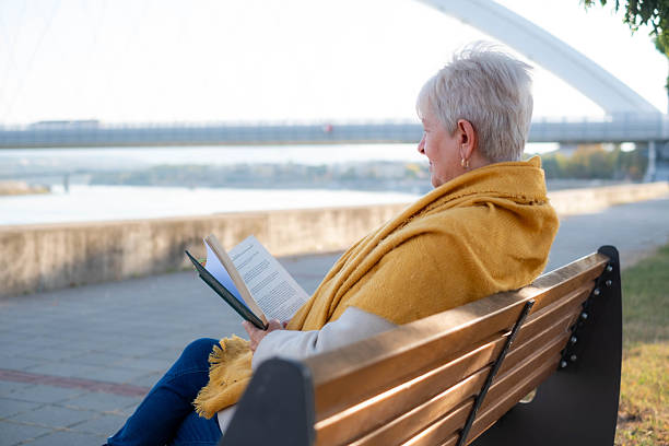 istock Senior woman reading book on park bench near river 2249053890