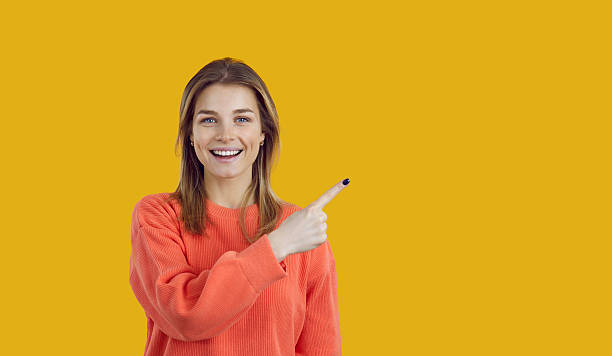 istock Happy woman on yellow background points finger sideways at copy space recommending offer or service. 2249067637