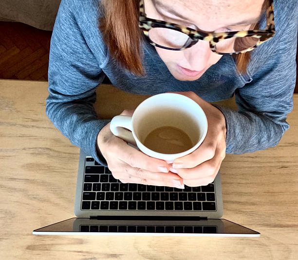 EX High angle CU woman holds coffee cup with both hands while looking at laptop computer screen in office