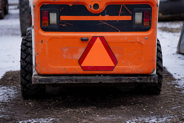 Rear view of an orange construction vehicle with reflective triangle sign, worn paint, rear lights and rugged tires standing on snowy ground in winter worksite conditions