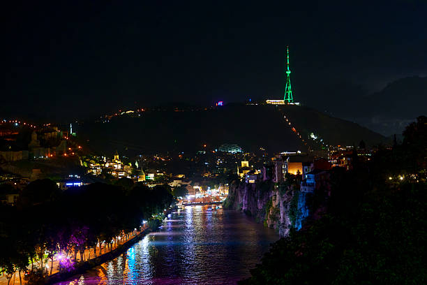 Colorful riverside lights reflect on Kura River while Mtatsminda TV tower glows green above Tbilisi.