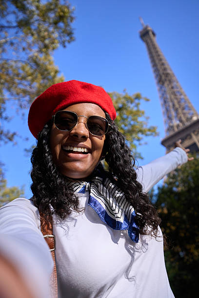 Vertical. Smiling African woman taking a selfie in Paris, pointing at the Eiffel Tower. Casual travel portrait with beret, sunglasses, conveying joy, energy and the excitement of sightseeing.
