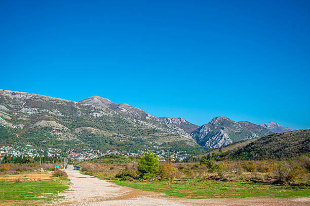 Gravel road leads through green fields toward the town of Sutomore, with clusters of houses at the foot of rocky mountains under a deep blue sky on a clear, sunny day in early autumn light!
