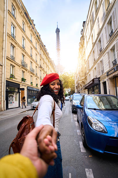 Vertical. Smiling African millennial woman holding hands and leading the way on a Paris street, Eiffel Tower in the background. Wide-angle travel portrait with lively city vibe and warm sunlight.