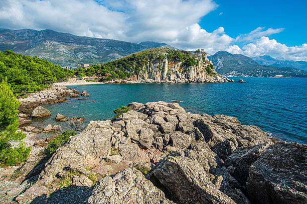 Rocky foreground overlooks a clear turquoise bay with scattered boulders, a small beach shelter, green pine trees, a steep coastal cliff, distant seaside town and layered mountains under sky