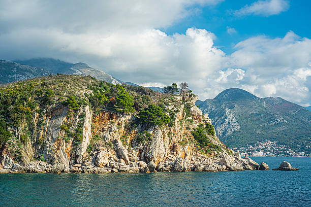 Sheer rocky cliffs covered with patches of green pine rise above calm blue sea, while distant mountains and a small coastal town appear beneath layered white clouds over the quiet horizon..