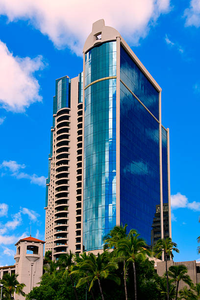 Exterior view of the Harbor Court building complex in downtown Honolulu, Oahu, Hawaii. This prominent postmodern mixed-use development features a taller residential condominium tower and a commercial office tower rising from a shared podium. The architecture showcases a distinctive stepped design with white concrete framing and reflective blue-green glass windows. Located adjacent to the Aloha Tower Marketplace and Honolulu Harbor, it stands as a key landmark in the city's urban waterfront skyline against a blue sky.