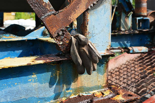 Worn work gloves resting on an aged, rusted metal framework with peeling blue paint. A scene of decay and industry, bathed in sunlight.