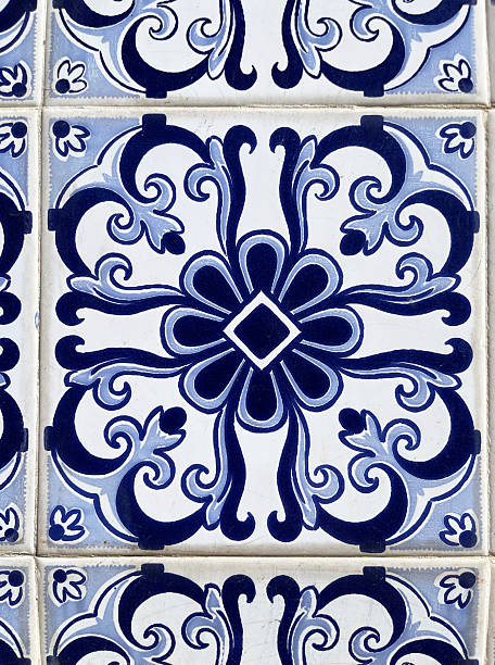Detail view of old, worn wall tiles that have a blue leaf abstract foliage design, with a flower design defining the center of a tile decorative pattern. The square tiles are positioned to have the surface pattern repeat and create an interconnected design from tile to tile. The plant motif and geometric visual on the smooth, glazed blue and white ceramic tile suggest an old-fashioned, early half of the 20th century aesthetic. These tiles are in Portugal, a country that decorates buildings with tile as an aspect of Portuguese culture.