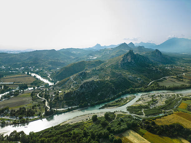 A drone view of a mountain range with rocky peaks, green hills and a winding river flowing through a wide valley under soft daylight.