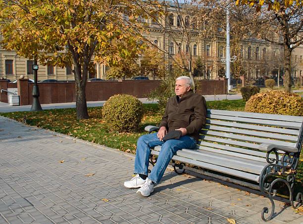 Mature elderly man sits alone on a wooden park bench during a sunny autumn day. He is dressed in a brown jacket, blue jeans, and white sneakers, holding a hat in his hands while calmly looking into the distance. The park is filled with yellow and orange fall foliage, trimmed bushes, and trees, with an elegant historic building visible in the background. This image is ideal for concepts related to retirement, peaceful lifestyle, solitude, senior health, mindfulness, autumn season, urban parks, and outdoor relaxation. Suitable for articles, advertisements, blogs, and campaigns about aging, wellness, and leisure.