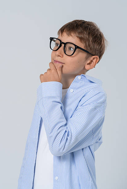 istock Thoughtful boy in glasses and striped shirt imagining ideas while posing with hand on chin on light background in a concept of curiosity and learning. 2249144990