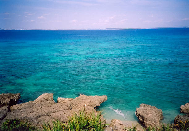 A clear tropical blue ocean viewed from a rocky coastline in Okinawa, Japan. The shallow turquoise water transitions into deep blue toward the horizon, creating a calm and vibrant seascape. Perfect for themes such as travel, tropical destinations, summer, nature backgrounds, and peaceful coastal scenery.