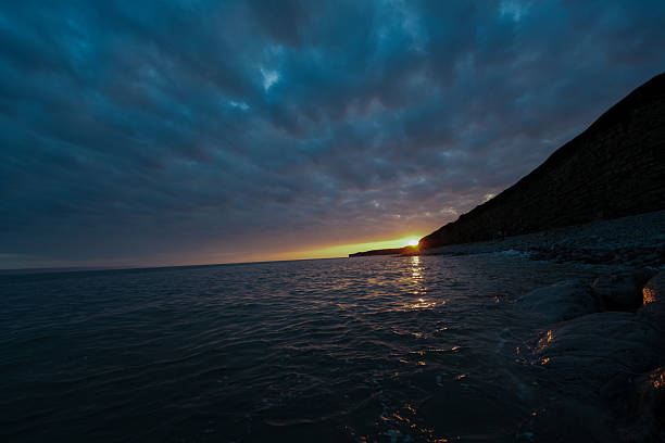 The sky is dark and cloudy, and the sun is setting over the ocean. The water is calm and still, and the rocks on the shoreline are illuminated by the sun's rays. The scene is serene and peaceful