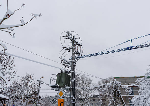 A tall power pole covered in snow rises against a pale winter sky, with cables stretching across the frame. The scene captures cold urban infrastructure during harsh seasonal weather and quiet snowy conditions.