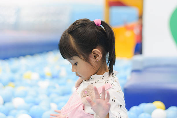 A close-up, candid profile shot of a young girl standing in a ball pit filled with light and dark blue balls. She has her hair pulled back and is looking down with a focused or contemplative expression, with one hand slightly raised. This intimate portrait captures a quiet moment amid the vibrant and busy activity of the indoor playground