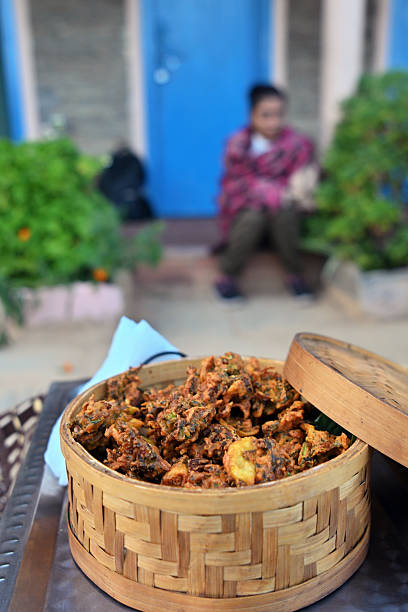 A rustic and homely scene featuring a basket of hot pakoras, with a casually seated woman blurred in the backdrop. Great for showcasing Indian tea-time snacks, countryside hospitality, cozy moments, and cultural food traditions.