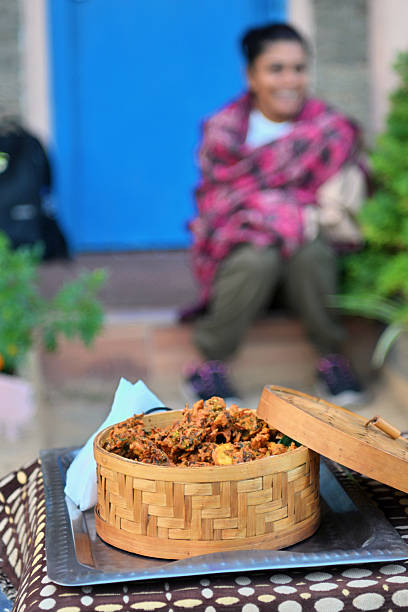 A close-up of freshly made vegetable pakoras served in a woven bamboo basket, placed on a tray. In the softly blurred background, a woman wrapped in a warm shawl sits outside a rustic cottage, creating a cozy and inviting homestay atmosphere. Indian cuisine, comfort food, rural living, and travel hospitality.