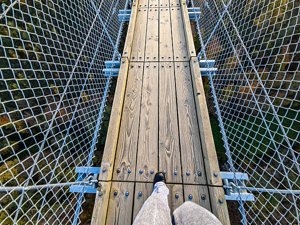 A solitary person walks across a long suspension bridge spanning a valley surrounded by autumn forest. The bright blue sky with white clouds contrasts sharply with the golden and orange foliage. The bridge seems endless, while the steel cables and wooden deck emphasize the height and adventure.