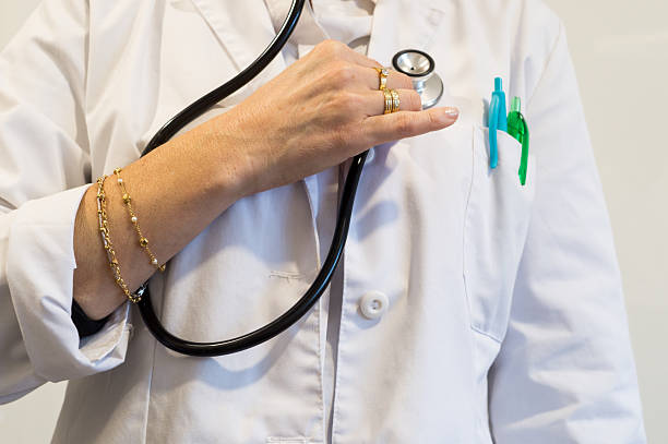 Mid-section view of a doctor (with pens in the pocket) adjusting a stethoscope, while clearly wearing both rings and a bracelet. Depicts a comprehensive failure to comply with medical cleanliness standards