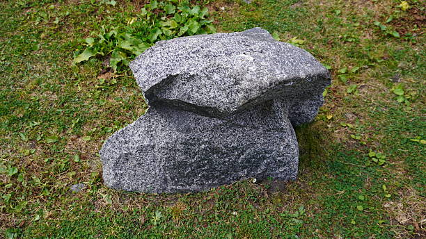 A large gray granite boulder sits on green grass with scattered yellow patches. The boulder has a rough, granular texture and is split into two distinct sections. The top section is slightly smaller and sits atop the larger base, creating a natural split in the stone. The boulder is weathered and has a matte finish. Small green plants and weeds grow around the base of the boulder. The grass is a mix of vibrant green and brown patches, indicating it may be late summer or early fall. The boulder takes up approximately one-third of the frame and is positioned slightly left of center. The lighting is natural and even, casting subtle shadows beneath the stone.