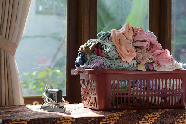A horizontal shot featuring an overflowing red-brown plastic laundry basket filled with clean clothes next to a small, old-fashioned electric iron, suggesting the next step in the chore: ironing. The items are placed on a patterned tablecloth by a large wooden window frame with white sheer curtains