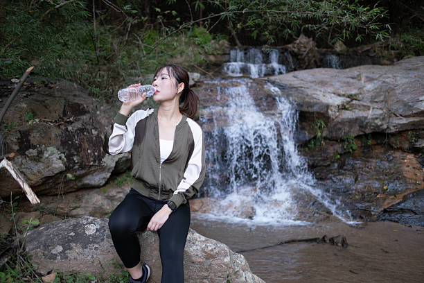 istock A woman sits on a rock near a waterfall, drinking water from a bottle 2249196412