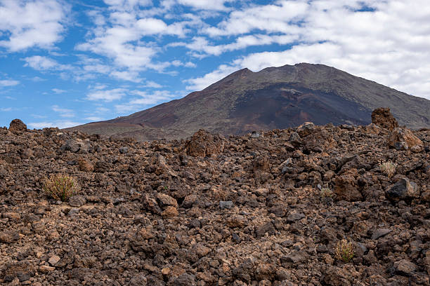 Rock formations and the rugged mountain slopes of El Teide National Park in Tenerife. The scene highlights layered lava structures, earthy tones, and the distinct summit of Teide under a deep blue sky — ideal for concepts of geology, wilderness, natural landscapes, volcanic environments, and conservation areas.