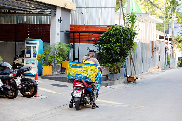 Thai motorcycle express delivery person with motorcycle is standing in front of local condominiums in residential district of Bangkok Chatuchak/Ladprao. Person has justy delivered something and has returnt back onto motorcycle and is checking mobile phone. On the back of motorcycle is a box. The box iswrapped with tapes. At left side some motorcycle are parked at condominium. Man is driving for FLASH Express
