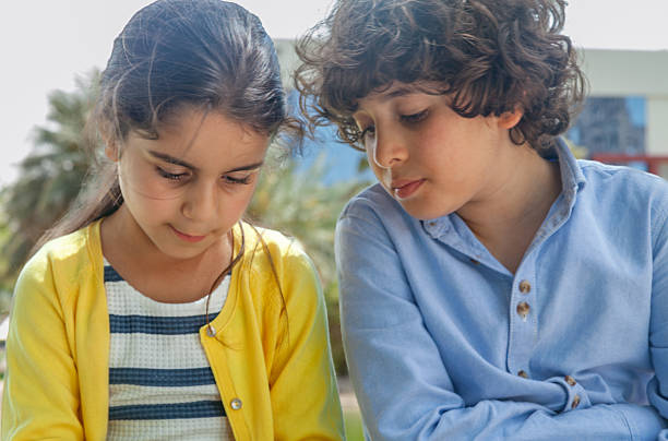 Two children sitting together on a bench in a lush park in Dubai, engrossed in a smartphone. The boy, dressed in a light blue shirt, and the girl, wearing a striped dress with a yellow cardigan, share a moment of concentration as they explore the device. The sunny weather, vibrant greenery, and palm trees create a peaceful and inviting backdrop, highlighting a blend of modern technology and natural surroundings