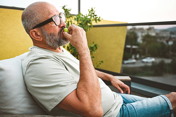 A mature adult man relaxes on a balcony and eats fruit.