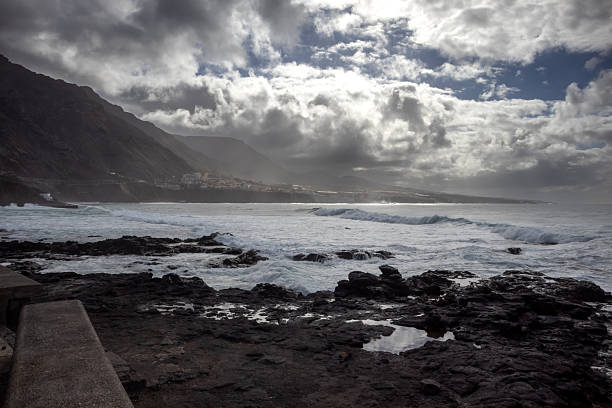 Natural coast made of volcanic black lava rocks. Atlantic ocean with waves in the autumn. Blue sky with intense clouds. Promenade in Punta de Hidalgo, Santa Cruz, Tenerife, Canary Islands, Spain.