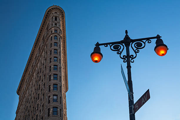 The iconic Flatiron building rises sharply toward a bright blue sky while a streetlamp adds urban charm in New York City during evening hours.