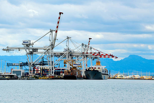 Honolulu, Oahu, Hawaii, USA: Matson Navigation Company container terminal at Pier 53 on Sand Island, Honolulu Harbor, Oahu. The image features the U.S.-flagged container ship R.J. Pfeiffer (IMO: 9002037) docked in the Kapalama Basin, being serviced by massive Ship-to-Shore (STS) gantry cranes. Built in 1992, this Jones Act vessel is a vital lifeline for Hawaii's supply chain. In the background, the lush slopes of the Koʻolau Mountain Range rise majestically above the industrial port, contrasting the heavy maritime infrastructure with the island's tropical landscape.