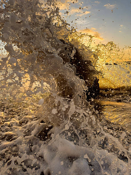 A high-impact, close-up photograph capturing a powerful wave cresting and splashing into motion on the wet, sandy shoreline at sunset. The low sun, positioned directly in the frame, casts a stunning golden and warm orange glow, backlighting the spray and creating sharp contrast and dramatic light refractions. The image evokes feelings of energy, tropical paradise, power, and the beauty of nature.