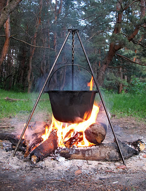 close-up of a black pot hanging from a tripod chain over a large campfire, surrounded by logs and trees in the evening forest