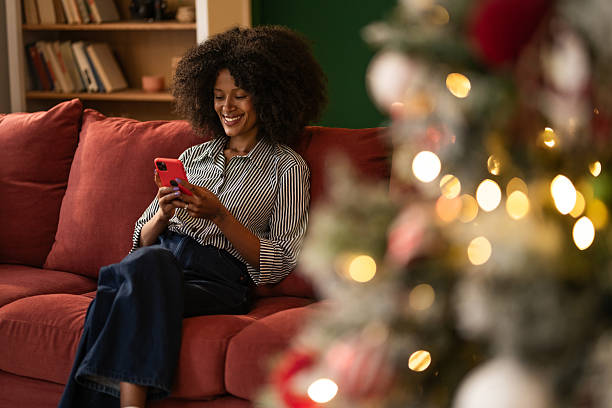 istock Young black woman using phone while sitting on cozy sofa at home on Christmas 2249240881