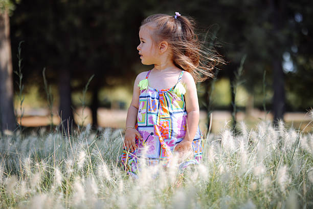 A pretty little girl is picking wildflowers in a lush meadow at sunset. She's alone in a peaceful natural setting, with a forest in the background.