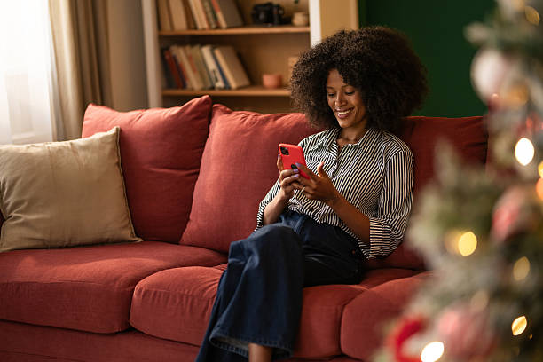 istock Young black woman using phone while sitting on cozy sofa at home on Christmas 2249240985
