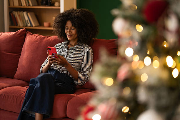 istock Young black woman using phone while sitting on cozy sofa at home on Christmas 2249241010