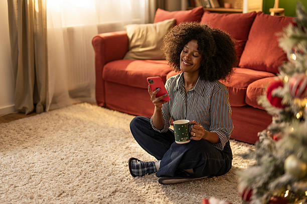 istock Young black woman using phone and enjoying a hot drink at home on Christmas 2249244431
