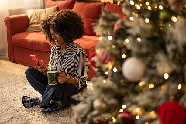 istock Young black woman using phone and enjoying a hot drink at home on Christmas 2249244579