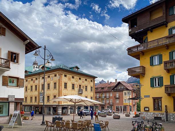 Cortina d'Ampezzo, Italy - June 8, 2022: Cityscape with Corso Italia and Piazza Roma; ornate facades with balconies and green shutters, cafes and shoppers under a blue sky with cloud in the Dolomites