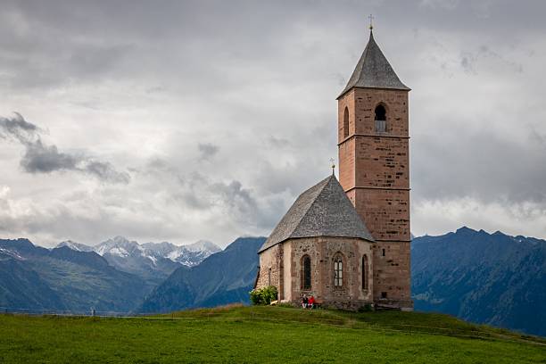 St. Kathrein Church (Chiesa di Santa Caterina) on a grassy hill in Hafling/Avelengo above Merano, infront of Oetztal alps mountains and against cloudy sky
