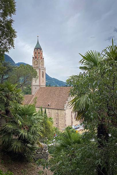 St. Nicholas Church in Merano, South Tyrol, Italy framed by palm trees; clock tower with green spire above rooftops, alpine hills behind under a sky with clouds