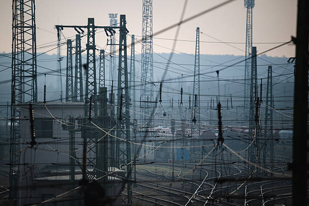 A hazy industrial landscape filled with numerous railway power line pylons and overhead cables, set against distant factory structures. The muted blue-gray tones and dense network of metal elements evoke a gritty, atmospheric industrial environment.