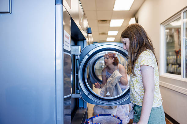 A young girl with long brown hair stands in front of an open washing machine at a laundromat. She is wearing a yellow patterned shirt and green pants, and appears to be in the process of loading laundry into the machine. The laundromat setting is visible with its rows of washing machines and bright fluorescent lighting. This scene captures a common household chore and the independence of youth learning life skills.