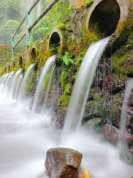 A scenic cascade pours through moss-covered stone arches, creating a misty, tranquil water feature amid vibrant green foliage. An ideal image for nature, travel, and outdoor lifestyle themes.