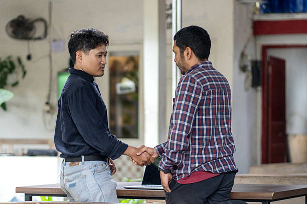Two Asian men shake hands beside a table with a laptop, suggesting an agreement or conclusion of a work discussion in an informal outdoor setting